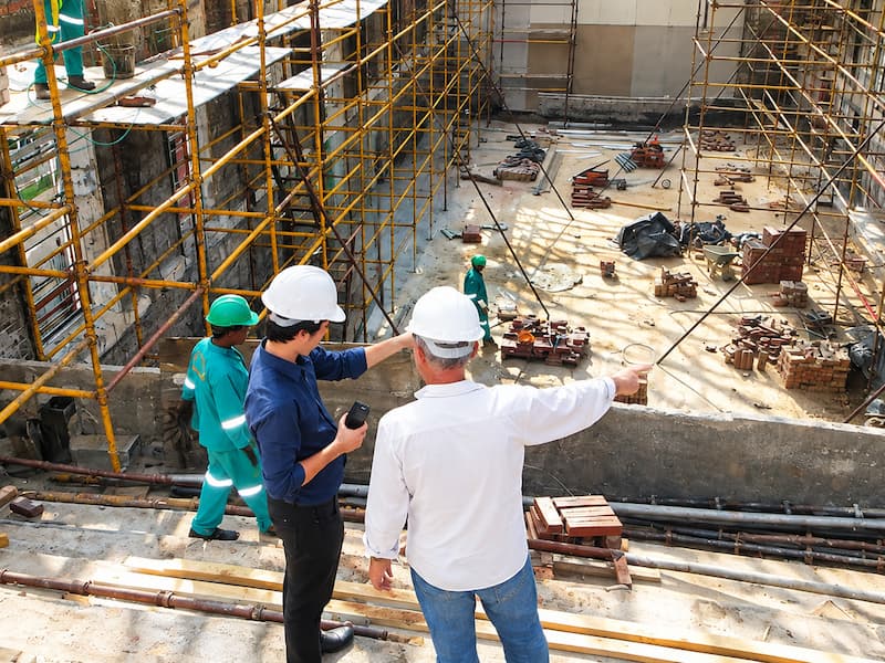 Construction supervisors overseeing workers and scaffolding at an active building site, with materials, tools, and concrete structures visible below.